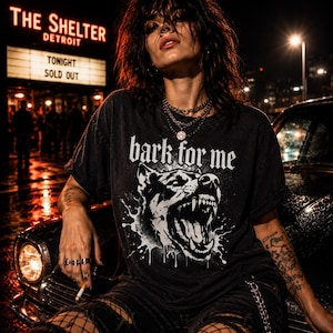 May include: Black graphic t-shirt with the text "bark for me" and a snarling dog illustration. The image also shows a marquee sign that reads "The Shelter Detroit Tonight Sold Out". The person is wearing multiple necklaces and rings.