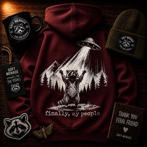 Op de afbeelding: Een bordeauxrode hoodie met een wasbeer-afbeelding die door een UFO wordt ontvoerd, met de tekst "finally, my people". Ook een zwarte mok, een groene beanie, een patch en een houten bord met "Thank You Feral Friend".