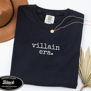 Black Comfort Colors 1717 tee with villain era in white Special Elite typewriter font. Cowboy hat and dried palm flat lay on white background. Evil queen dark humor graphic tee.