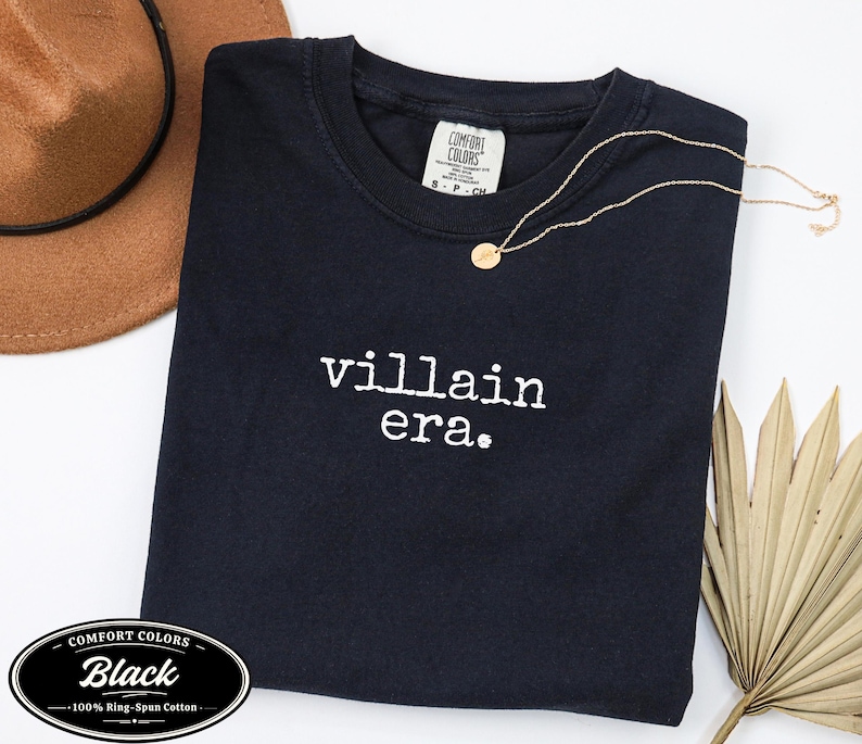 Black Comfort Colors 1717 tee with villain era in white Special Elite typewriter font. Cowboy hat and dried palm flat lay on white background. Evil queen dark humor graphic tee.