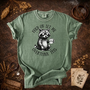 Hold On Let Me Overthink This shirt in Moss — Comfort Colors 1717 raccoon graphic tee — earthy green colorway flat lay — funny women’s tshirt — panic raccoon holding coffee mug design