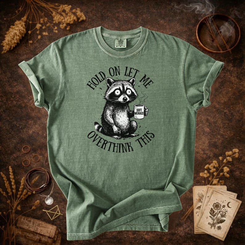 Hold On Let Me Overthink This shirt in Moss — Comfort Colors 1717 raccoon graphic tee — earthy green colorway flat lay — funny women’s tshirt — panic raccoon holding coffee mug design
