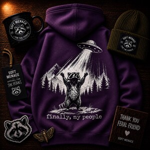 Op de afbeelding: Een paarse hoodie met een wasbeer die door een UFO wordt ontvoerd, met de tekst "finally, my people". Ook afgebeeld zijn een zwarte mok, een groene beanie, een label, een sticker en een houten bord, allemaal met het "Soft Menace"-logo.