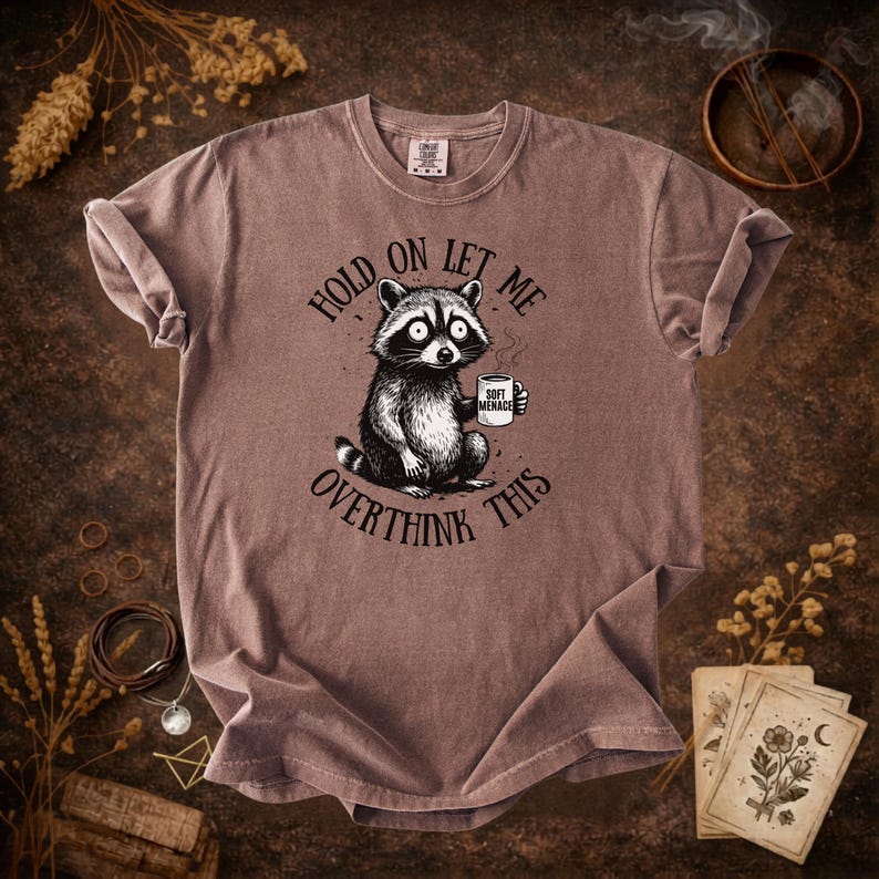 May include: A mauve-colored t-shirt featuring a raccoon holding a mug with the words "Soft Menace." The shirt has the text "Hold on let me overthink this" around the raccoon. The shirt is a comfortable, casual wear item.