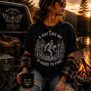 以下が含まれることがあります： 「A Guy Like Me Is Hard To Find」のテキストとビッグフットのグラフィックがプリントされた黒いTシャツ。画像には、アライグマのデザインの黒いマグカップ、ヴィンテージスタイルのトランク、「Soft Menace Co.」のロゴも表示されています。