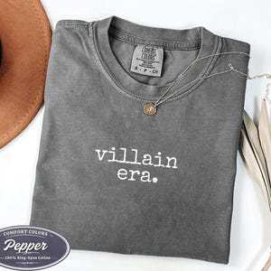 Pepper Comfort Colors 1717 tee with villain era in white Special Elite typewriter font. Cowboy hat and dried palm flat lay on white background. Dark academia evil queen shirt in heathered gray