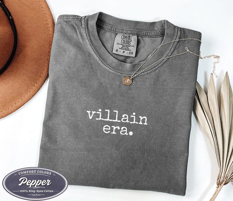 Pepper Comfort Colors 1717 tee with villain era in white Special Elite typewriter font. Cowboy hat and dried palm flat lay on white background. Dark academia evil queen shirt in heathered gray