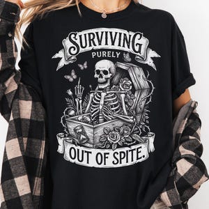May include: Black graphic t-shirt with the text "SURVIVING PURELY OUT OF SPITE." The design features a skeleton in a coffin, surrounded by roses and butterflies. The skeleton is giving the middle finger.