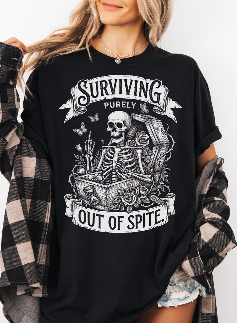 May include: Black graphic t-shirt with the text "SURVIVING PURELY OUT OF SPITE." The design features a skeleton in a coffin, surrounded by roses and butterflies. The skeleton is giving the middle finger.