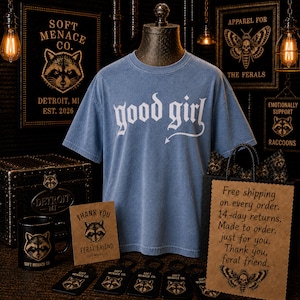 Blue Jean Comfort Colors 1717 “good girl” graphic t-shirt displayed on vintage mannequin in dark gothic boutique setting. Warm cinematic lighting, raccoon branding details, and alternative punk decor create moody editorial fashion scene. Soft Menace Co. women’s streetwear graphic tee featuring distressed gothic typography, relaxed unisex fit, garment dyed cotton texture, and edgy dark feminine clothing aesthetic.