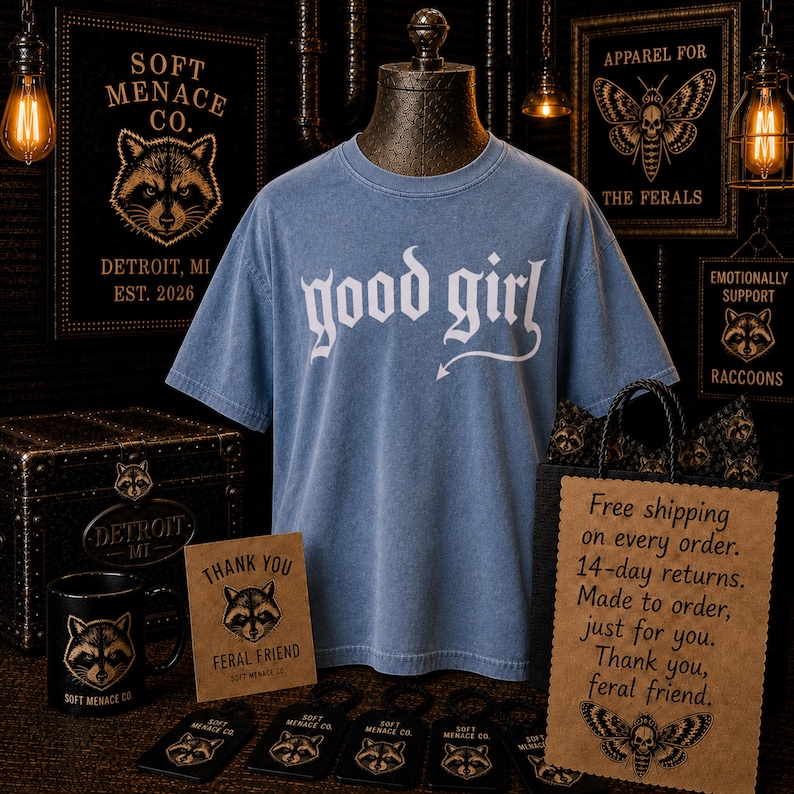 Blue Jean Comfort Colors 1717 “good girl” graphic t-shirt displayed on vintage mannequin in dark gothic boutique setting. Warm cinematic lighting, raccoon branding details, and alternative punk decor create moody editorial fashion scene. Soft Menace Co. women’s streetwear graphic tee featuring distressed gothic typography, relaxed unisex fit, garment dyed cotton texture, and edgy dark feminine clothing aesthetic.