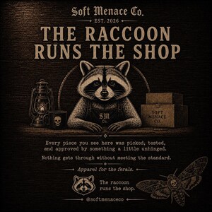 Op de afbeelding: Illustratie met een wasbeer met een schort, met de tekst "THE RACCOON RUNS THE SHOP." Andere elementen zijn een lantaarn, een mok met een schedel en dozen. De tekst "Soft Menace Co. EST. 2026" is ook aanwezig. Apparel for the ferals.