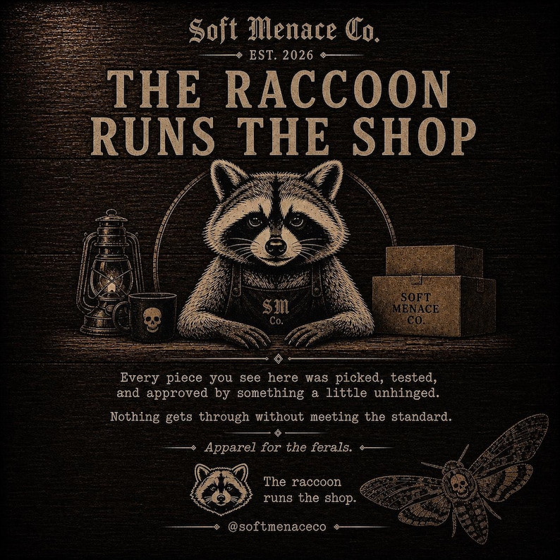 Op de afbeelding: Illustratie met een wasbeer met een schort, met de tekst "THE RACCOON RUNS THE SHOP." Andere elementen zijn een lantaarn, een mok met een schedel en dozen. De tekst "Soft Menace Co. EST. 2026" is ook aanwezig. Apparel for the ferals.