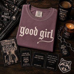 Berry purple Comfort Colors 1717 “good girl” gothic t-shirt folded on dark wood flat lay with cinematic amber lighting. Includes Soft Menace Co. punk props, chains, stickers, and dark feminine aesthetic styling. Vintage garment dyed alternative graphic tee with distressed cream blackletter typography designed for gothic fashion lovers, punk streetwear shoppers, and edgy women’s casual apparel collections.