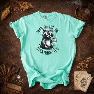 May include: A light turquoise t-shirt featuring a raccoon holding a coffee cup. The raccoon is illustrated in black and white with the text "HOLD ON LET ME OVERTHINK THIS". The coffee cup reads "SOFT MENACE".