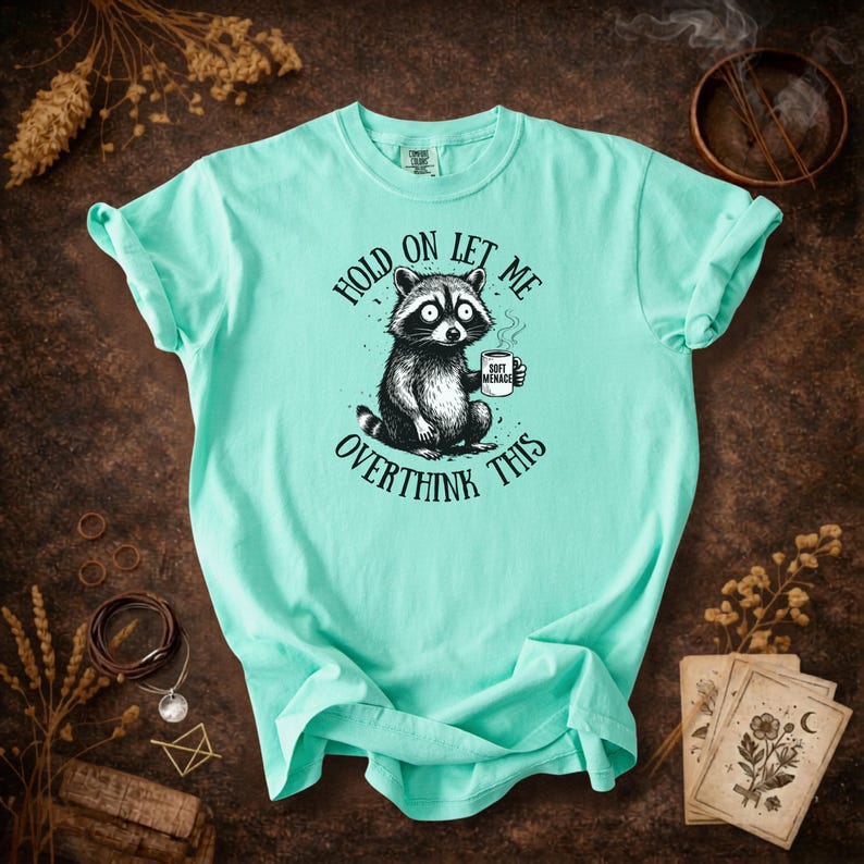 May include: A light turquoise t-shirt featuring a raccoon holding a coffee cup. The raccoon is illustrated in black and white with the text "HOLD ON LET ME OVERTHINK THIS". The coffee cup reads "SOFT MENACE".