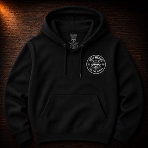 Op de afbeelding: Zwarte hoodie met een kangoeroezak en trekkoord. De voorkant heeft een cirkelvormige afbeelding met een wasbeerillustratie en de tekst "SOFT MENACE" en "FOR THE FERALS". De hoodie heeft een label op de capuchon.