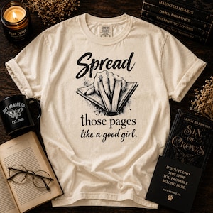 Op de afbeelding: Een crèmekleurig T-shirt met korte mouwen en een zwarte afbeelding van een hand die op een open boek rust. De tekst luidt: "Spread those pages like a good girl." Het shirt heeft een ronde hals en een Comfort Colors-label in de binnenkraag.
