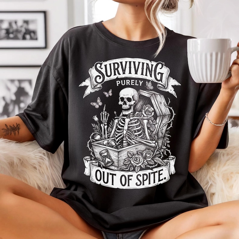 Black skeleton shirt on woman holding white coffee mug, fluffy blanket background. Dark humor gothic graphic tee on Comfort Colors 1717. Surviving Purely Out Of Spite — funny dark academia shirt.