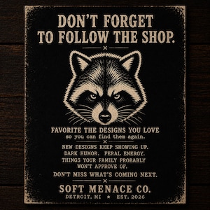 Soft Menace Co. follow shop reminder card featuring raccoon mascot artwork, gothic typography, and dark alternative branding design. Cinematic amber lighting over dark walnut wood background with punk-inspired details and moody editorial aesthetic. Encourages shoppers to favorite the Etsy shop, follow for new releases, and stay updated on future gothic graphic tees, dark feminine apparel, punk streetwear, and alternative fashion collections.