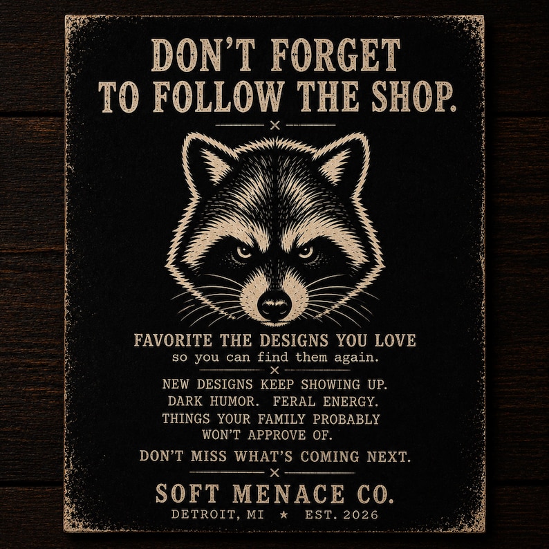 Soft Menace Co. follow shop reminder card featuring raccoon mascot artwork, gothic typography, and dark alternative branding design. Cinematic amber lighting over dark walnut wood background with punk-inspired details and moody editorial aesthetic. Encourages shoppers to favorite the Etsy shop, follow for new releases, and stay updated on future gothic graphic tees, dark feminine apparel, punk streetwear, and alternative fashion collections.