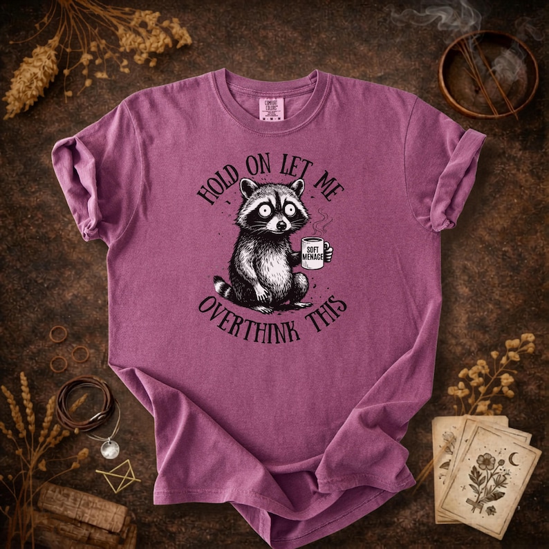 May include: A mauve t-shirt featuring a raccoon illustration holding a mug with the words "Soft Menace." The text "Hold on let me overthink this" is printed around the raccoon. The shirt has a crew neck and short sleeves.