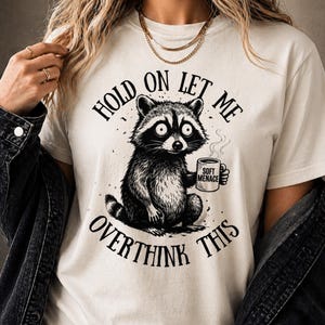 Cream-colored t-shirt featuring a detailed black and white illustration of a raccoon holding a mug. The text on the shirt reads "HOLD ON LET ME OVERTHINK THIS" and "SOFT MENACE" on the mug.