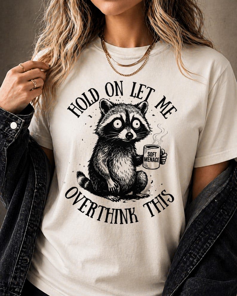 Cream-colored t-shirt featuring a detailed black and white illustration of a raccoon holding a mug. The text on the shirt reads "HOLD ON LET ME OVERTHINK THIS" and "SOFT MENACE" on the mug.