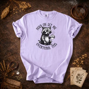 May include: A light purple t-shirt featuring a raccoon holding a coffee mug. The raccoon is black and white with large eyes. Text on the shirt reads "Hold on let me overthink this" and "Soft Menace".