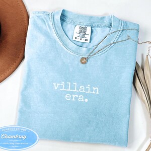 May include: A light blue t-shirt with the words "villain era." printed in white. The shirt has a crew neck and short sleeves. A silver necklace with a pendant rests on the shirt. The shirt is made of 100% ring-spun cotton.