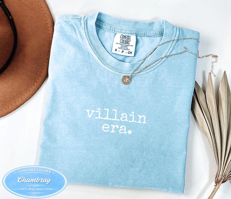 May include: A light blue t-shirt with the words "villain era." printed in white. The shirt has a crew neck and short sleeves. A silver necklace with a pendant rests on the shirt. The shirt is made of 100% ring-spun cotton.