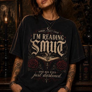 May include: A charcoal black short-sleeve t-shirt featuring a graphic print with the text SORRY, CAN'T TALK I'M READING SMUT AND HIS EYES just darkened. The design includes an open book, a pair of eyes, and two red roses with dark green leaves.