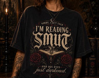 May include: A charcoal black short-sleeve t-shirt featuring a graphic print with the text SORRY, CAN'T TALK I'M READING SMUT AND HIS EYES just darkened. The design includes an open book, a pair of eyes, and two red roses with dark green leaves.