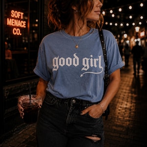 Woman wearing faded blue Comfort Colors 1717 “good girl” gothic graphic t-shirt with distressed cream blackletter typography. Alternative streetwear aesthetic with layered necklaces, dark jeans, moody lighting, and casual punk styling. Soft Menace Co. graphic tee lifestyle image featuring dark feminine fashion, edgy aesthetic clothing, gothic typography apparel, and relaxed oversized vintage garment dyed t-shirt vibes.