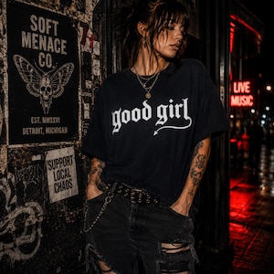 Alternative woman wearing a black Comfort Colors 1717 graphic t-shirt with “good girl” gothic lettering in distressed cream print. Moody nightlife street scene with red neon glow, tattoos, layered necklaces, ripped black jeans, and dark punk aesthetic. Soft Menace Co. dark feminine apparel photo with cinematic lighting, edgy streetwear styling, and gothic fashion energy designed for women who love alternative clothing and dark humor fashion.