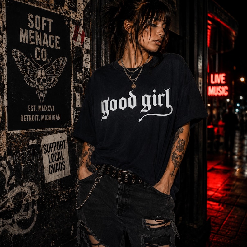 Alternative woman wearing a black Comfort Colors 1717 graphic t-shirt with “good girl” gothic lettering in distressed cream print. Moody nightlife street scene with red neon glow, tattoos, layered necklaces, ripped black jeans, and dark punk aesthetic. Soft Menace Co. dark feminine apparel photo with cinematic lighting, edgy streetwear styling, and gothic fashion energy designed for women who love alternative clothing and dark humor fashion.