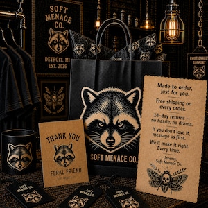 Soft Menace Co. branded packaging and thank you insert display featuring raccoon mascot artwork, gothic branding cards, apparel packaging materials, stickers, and premium small business presentation. Dark walnut wood background with cinematic amber lighting and punk-inspired props. Alternative clothing brand packaging aesthetic for gothic apparel shoppers and dark feminine streetwear customers.