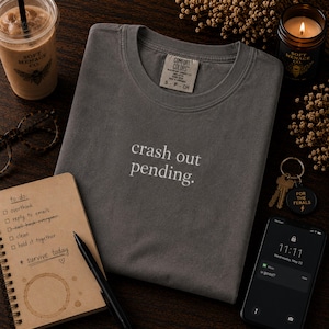Pode incluir: Uma camiseta cinza Comfort Colors com a frase "crash out pending" em branco. A imagem inclui uma lista de tarefas, um telefone exibindo uma mensagem, uma vela, uma bebida de café e chaves.