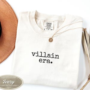 Ivory Comfort Colors 1717 tee with villain era in black Special Elite typewriter font. Folded flat lay with brown cowboy hat, gold necklace, and dried palm leaf on white background. Trendy unhinged graphic tee.