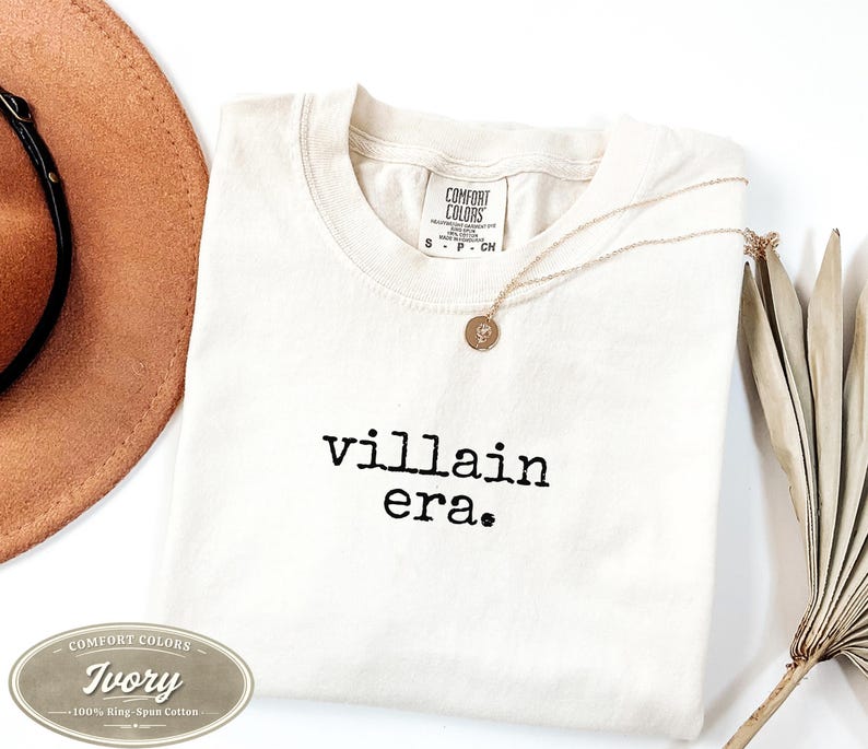 Ivory Comfort Colors 1717 tee with villain era in black Special Elite typewriter font. Folded flat lay with brown cowboy hat, gold necklace, and dried palm leaf on white background. Trendy unhinged graphic tee.