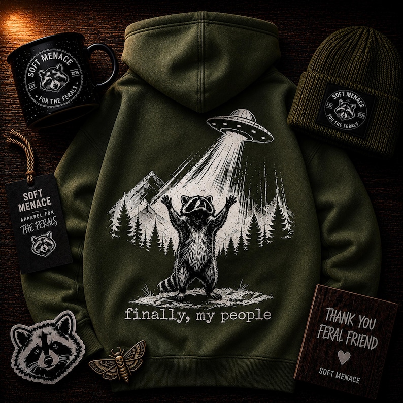 Op de afbeelding: Een olijfgroene hoodie met een afbeelding van een wasbeer die door een UFO wordt opgetild, met de tekst "finally, my people". Ook afgebeeld zijn een zwarte mok, muts, patches en een houten bord, allemaal met het "Soft Menace" logo.
