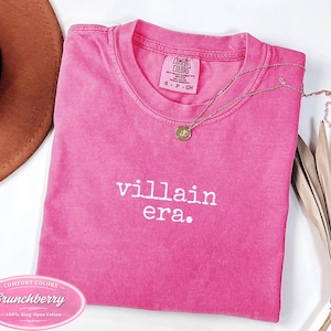 Crunchberry Comfort Colors 1717 tee with villain era in white Special Elite typewriter font. Cowboy hat and dried palm flat lay styling on white background. Dark humor evil queen shirt in bold pink.