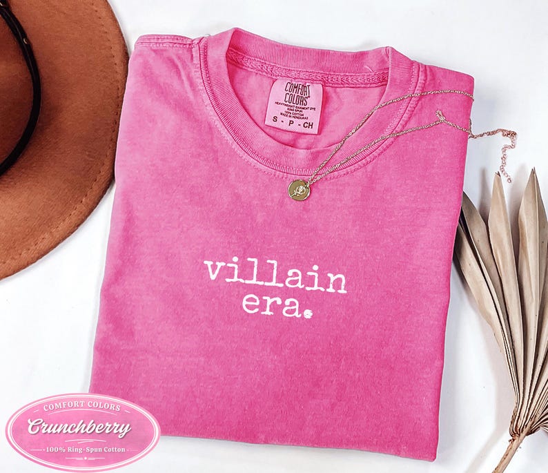 Crunchberry Comfort Colors 1717 tee with villain era in white Special Elite typewriter font. Cowboy hat and dried palm flat lay styling on white background. Dark humor evil queen shirt in bold pink.