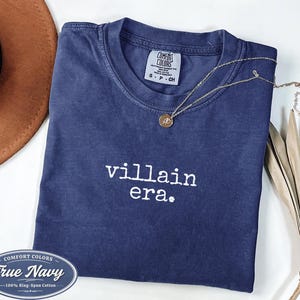 True Navy Comfort Colors 1717 tee with villain era in white Special Elite typewriter font. Folded flat lay with brown cowboy hat, gold necklace, and dried palm on white background. Dark academia goth shirt