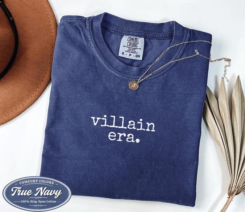 True Navy Comfort Colors 1717 tee with villain era in white Special Elite typewriter font. Folded flat lay with brown cowboy hat, gold necklace, and dried palm on white background. Dark academia goth shirt
