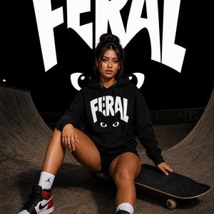 May include: A black hooded sweatshirt featuring the word FERAL in white capital letters across the chest, positioned above a graphic of two white cat eyes. The hoodie includes a front kangaroo pocket and long sleeves with ribbed cuffs.