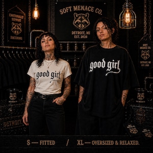 Two models wearing Soft Menace Co. “good girl” gothic graphic t-shirts in fitted and oversized styling examples. Alternative fashion fit comparison showing relaxed unisex Comfort Colors 1717 sizing on women with dark punk aesthetic and cinematic boutique lighting. Graphic tee styling reference for gothic streetwear shoppers wanting fitted or oversized dark feminine clothing looks.