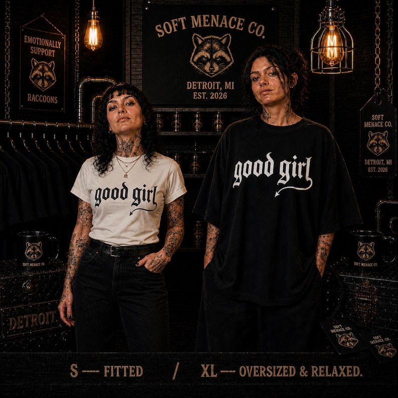 Two models wearing Soft Menace Co. “good girl” gothic graphic t-shirts in fitted and oversized styling examples. Alternative fashion fit comparison showing relaxed unisex Comfort Colors 1717 sizing on women with dark punk aesthetic and cinematic boutique lighting. Graphic tee styling reference for gothic streetwear shoppers wanting fitted or oversized dark feminine clothing looks.