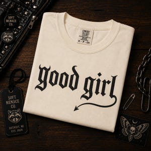 Folded ivory Comfort Colors 1717 graphic t-shirt with “good girl” gothic blackletter design displayed on dark walnut wood surface. Cinematic warm lighting with punk leather jacket, chains, raccoon branding tag, and dark alternative props. Soft Menace Co. premium flat lay mockup showcasing vintage garment dyed tee texture, gothic streetwear aesthetic, and moody punk-inspired apparel branding.