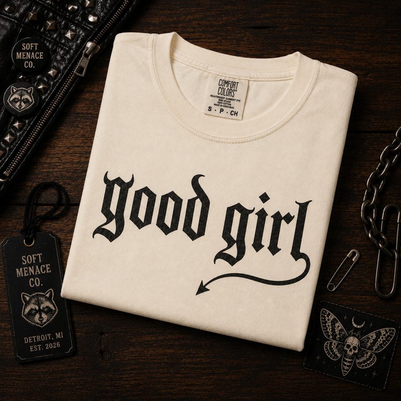 Folded ivory Comfort Colors 1717 graphic t-shirt with “good girl” gothic blackletter design displayed on dark walnut wood surface. Cinematic warm lighting with punk leather jacket, chains, raccoon branding tag, and dark alternative props. Soft Menace Co. premium flat lay mockup showcasing vintage garment dyed tee texture, gothic streetwear aesthetic, and moody punk-inspired apparel branding.